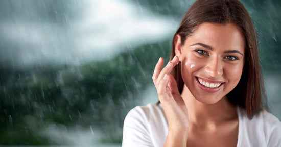 A smiling woman applies face cream to her cheek while standing in the rain, with a blurred green and grey background suggesting a natural, outdoor setting. She has straight brown hair and is wearing a white top