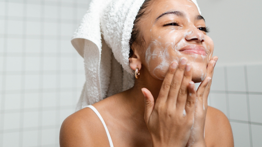 A woman with a white towel wrapped around her head is smiling while gently washing her face with a foaming cleanser in a tiled bathroom.