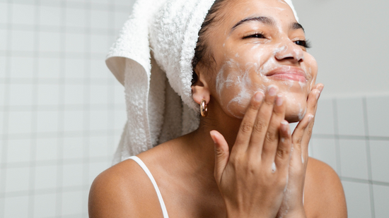 A woman with a white towel wrapped around her head is smiling while gently washing her face with a foaming cleanser in a tiled bathroom.