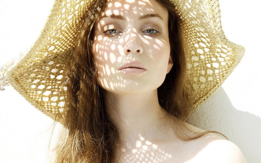 A woman wearing a straw hat with sunlight casting patterned shadows on her face, representing summer skincare and protection