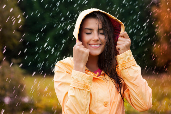 A young woman in a yellow raincoat standing in the rain. Her wet hair peeks out from under the hood as raindrops fall around her. The background features lush greenery and warm autumn tones, creating a vibrant and refreshing monsoon atmosphere.