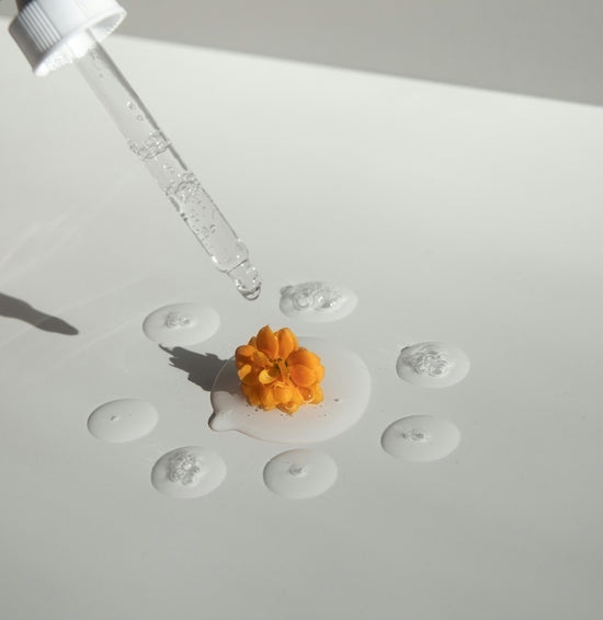 A glass serum dropper dispensing a clear skincare serum onto a white surface with an orange marigold flower placed at the center, surrounded by small serum droplets. The aesthetic is clean, minimal, and natural, reflecting purity and skincare.